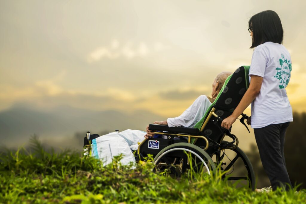 dziewczyna w krótkich, ciemnych włosach stoi za wózkiem inwalidzkim, na którym siedzi starszy człowiek. Obie osoby są skierowane bokiem. stoją na zielonej trawiea w tle widać zachód słońca